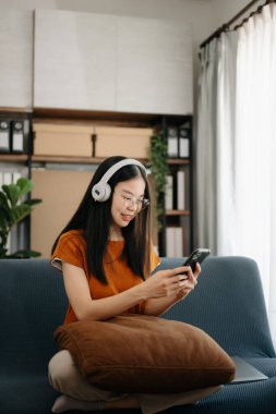 Çekici, mutlu, Asyalı genç bir öğrenci üniversite kütüphanesinde okuyor, koltukta oturuyor, kulaklık ve akıllı telefon kullanıyor, görüntülü konuşma yapıyor..