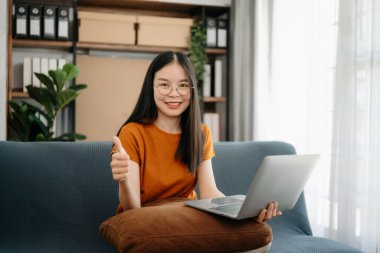  Asyalı kız kütüphanesi kanepede dizüstü bilgisayar ve tabletle internette arama yapıyor. 