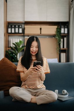 Asyalı kadın evdeki kanepede akıllı telefon kullanıyor.