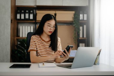 İş kadını sabah ışığında ofis masasında akıllı telefon ve laptop kullanıyor.