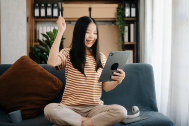 Çekici, mutlu Asyalı öğrenci üniversite kütüphanesinde okuyor, kanepede oturuyor, dijital tablet bilgisayar kullanıyor.