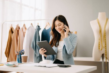 Asyalı terzi kadın terzi atölyesinde kıyafetlerle çalışıyor. Stüdyoda güzel genç bayan moda tasarımcısı