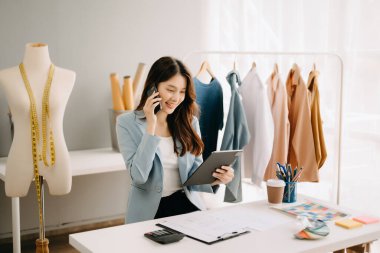 Asyalı terzi kadın terzi atölyesinde kıyafetlerle çalışıyor. Stüdyoda güzel genç bayan moda tasarımcısı
