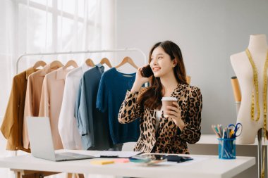 Asyalı terzi kadın terzi atölyesinde kıyafetlerle çalışıyor. Stüdyoda güzel genç bayan moda tasarımcısı