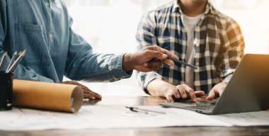 İki meslektaş dizüstü bilgisayar ve planlarla çalışan mimari projeleri tartışıyor 