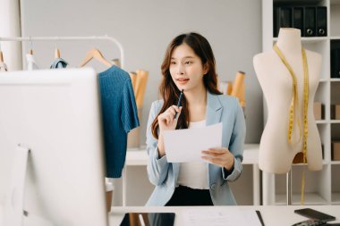 Asyalı terzi kadın terzi atölyesinde kıyafetlerle çalışıyor. Stüdyoda güzel genç bayan moda tasarımcısı