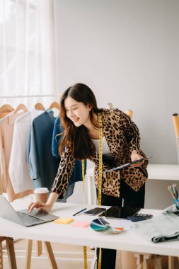 Asyalı terzi kadın terzi atölyesinde kıyafetlerle çalışıyor. Stüdyoda güzel genç bayan moda tasarımcısı