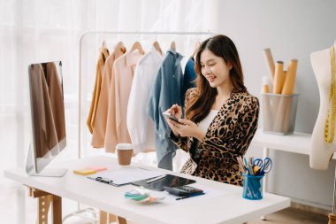 Asyalı terzi kadın terzi atölyesinde kıyafetlerle çalışıyor. Stüdyoda güzel genç bayan moda tasarımcısı