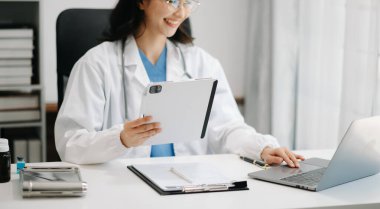 Güzel doktor modern ofis hastanesinde masada oturuyor. Çekici terapist gülümsemesi ve dijital tablet ve dizüstü bilgisayar kullanımı