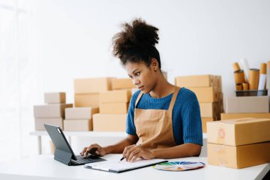 Başlangıçta küçük işletme KOBİ 'si, girişimci sahibi Afrikalı kadın tablet bilgisayar kullanıyor, paket ürün kutusu hazırlamak için online alışveriş siparişi alıyor ve kontrol ediyor.