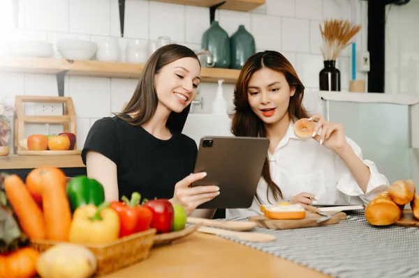 stock image Two beautiful women in the kitchen in an apron, fresh vegetables on the table, writes down her favourite recipes, comes up with ideas for dishes 