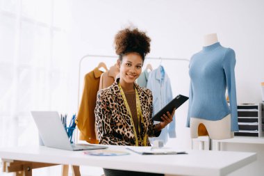 Sakin, kıvırcık esmer, koyu tenli kadın modern moda tasarımcısının ofisinde masanın üstünde ve elinde tablet var.  