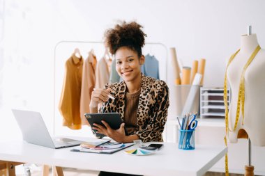 Sakin, kıvırcık esmer, koyu tenli kadın modern moda tasarımcısının ofisinde masanın üstünde ve elinde tablet var.  