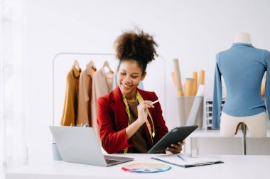 Sakin, kıvırcık esmer, koyu tenli kadın modern moda tasarımcısının ofisinde masanın üstünde ve elinde tablet var.  