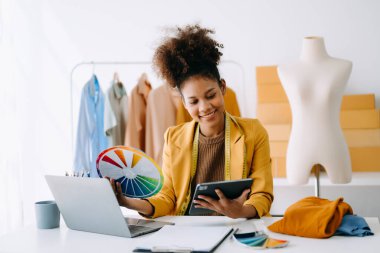 Sakin, kıvırcık esmer, koyu tenli kadın modern moda tasarımcısının ofisinde masanın üstünde ve elinde tablet var.  