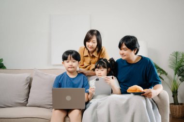 Asyalı ebeveynler ve çocuklar koltukta otururken birlikte cihaz kullanıyorlar. Teknoloji bağımlısı aile evde eğleniyor.