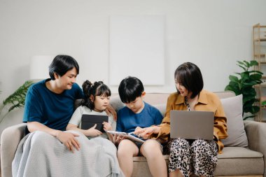 Asyalı ebeveynler ve çocuklar koltukta otururken birlikte cihaz kullanıyorlar. Teknoloji bağımlısı aile evde eğleniyor.