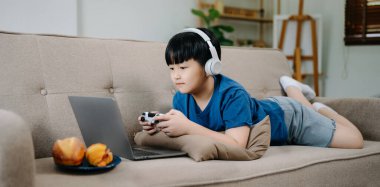 Genç Asyalı çocuk evde kulaklık takıp kanepede yatarken joystick ile video oyunu oynuyor. 