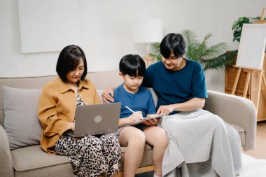 Asyalı anne ve baba ve oğul koltukta otururken birlikte cihaz kullanıyorlar. Teknoloji bağımlısı aile evde eğleniyor.