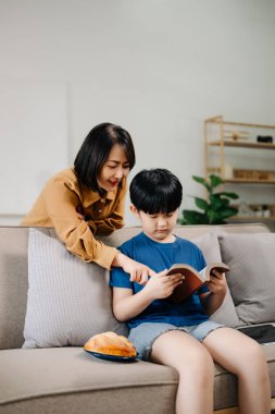 Mutlu Asyalı anne ve oğlu aile kanepesinde oturup birlikte kitap okuyorlar.