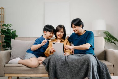 Asyalı anne ve baba ve oğul koltukta otururken birlikte cihaz kullanıyorlar. Teknoloji bağımlısı aile evde eğleniyor.