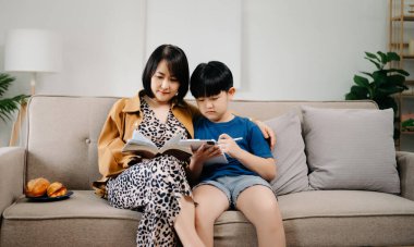 Mutlu Asyalı anne ve oğlu aile kanepesinde oturup birlikte kitap okuyorlar.