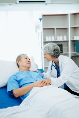 Beyaz takım elbiseli Asyalı doktor hastanede yatağında yatan yaşlı bir hastayla konuşuyor. 