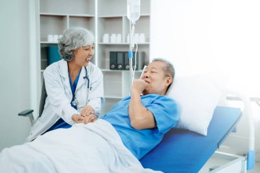 Beyaz takım elbiseli Asyalı doktor hastanede yatağında yatan yaşlı bir hastayla konuşuyor. 
