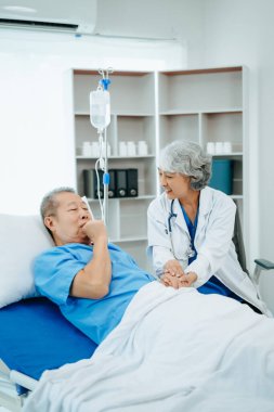 Beyaz takım elbiseli Asyalı doktor hastanede yatağında yatan yaşlı bir hastayla konuşuyor. 
