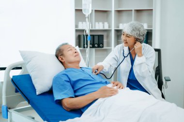Beyaz takım elbiseli Asyalı doktor hastanede yatağında yatan yaşlı bir hastayla konuşuyor. 