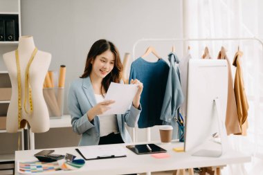 Asyalı terzi kadın terzi atölyesinde giysiler üzerinde çalışıyor. Güzel genç bayan moda tasarımcısı gülümsemesi ve stüdyo başarısından sonra