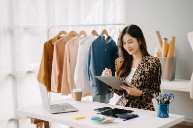 Asyalı terzi kadın terzi atölyesinde giysiler üzerinde çalışıyor. Güzel genç bayan moda tasarımcısı gülümsemesi ve stüdyo başarısından sonra