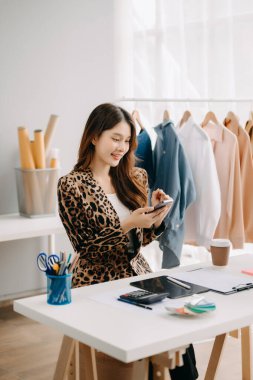 Asyalı terzi kadın terzi atölyesinde giysiler üzerinde çalışıyor. Güzel genç bayan moda tasarımcısı gülümsemesi ve stüdyo başarısından sonra