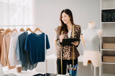 Asyalı terzi kadın terzi atölyesinde giysiler üzerinde çalışıyor. Güzel genç bayan moda tasarımcısı gülümsemesi ve stüdyo başarısından sonra