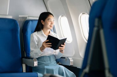 Genç Asyalı yönetici birinci sınıfta, not defteriyle birden fazla iş yapıyor. Stil içinde seyahat et, zarafetle çalış..