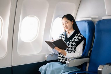 Genç Asyalı yönetici birinci sınıfta, not defteriyle birden fazla iş yapıyor. Stil içinde seyahat et, zarafetle çalış..