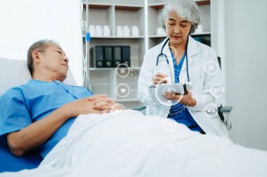 Beyaz takım elbiseli Asyalı doktor hastanede yatağında yatan yaşlı bir hastayla konuşuyor. 