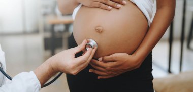 Pregnant African woman has appointment with doctor at clinic. Male specialist with stethoscope listens to baby's heartbeat in mother's belly. Pregnancy, health care concept clipart