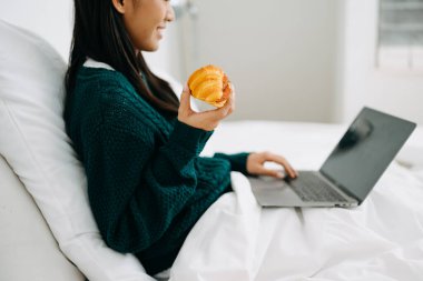 Beyaz bir yatakta, elinde kahve fincanı ve evinde dizüstü bilgisayar kullanan güzel bir genç kadın.