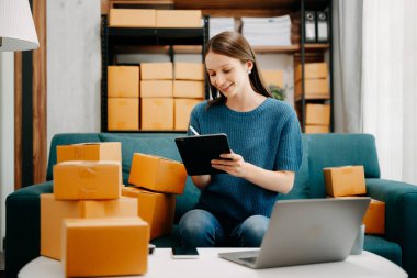 Genç işyeri sahibi kadın, dijital tabletteki müşterilere teslimat için paket kutusu hazırlıyor ve online siparişleri kontrol ediyor. İnternetten alışveriş konsepti. Ofisteki kanepede.