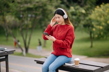 Güzel, genç Asyalı bir kadın kulaklıkla müzik dinliyor. Mutlu ve rahat, gülen yüzlü. Sabah parkta.