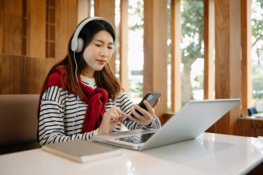 Attractive happy young Asian student studying at the college library, sitting at the desk, using a laptop computer and headphones having a video chat