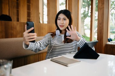 Üniversite kütüphanesinde okuyan, akıllı telefon, dijital tablet ve kulaklık kullanan çekici, mutlu, Asyalı bir öğrenci.