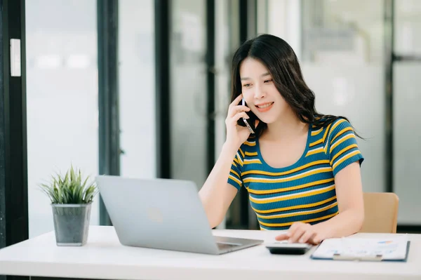 Smiling Asian woman talking on the phone with a customer. Young positive female accountant using smartphone talking to team at her desk