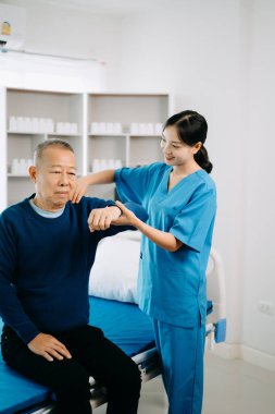Asyalı fizyoterapist, hastanede egzersiz sırasında erkek hastanın kolunu esnetmesine yardım ediyor. Yaşlı adamın sırt sorunları var..