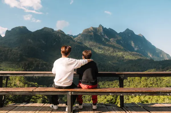 Rear view image of two male travelers sitting and looking at a beautiful mountain, sky field and nature view in morning light