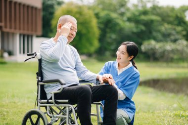 Tekerlekli sandalyede baş ağrısı olan yaşlı Asyalı son sınıf öğrencisi ve Asyalı dikkatli bir bakıcı.. 