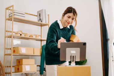 Genç bayan iş sahibi dizüstü bilgisayar kullanıyor ve evdeki müşterilere çevrimiçi siparişler göndermek için paketler hazırlıyor. Çevrimiçi Alışveriş kavramı