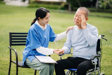 Tekerlekli sandalyede baş ağrısı olan yaşlı Asyalı yaşlı bir adam ve hastane bahçesinde dosyası olan Asyalı dikkatli bir bakıcı.