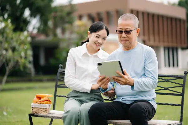 Tekerlekli sandalyedeki yaşlı Asyalı son sınıf öğrencisi Asyalı dikkatli bir bakıcı tabletle ve hastane bahçesi konseptiyle yemek yiyor..